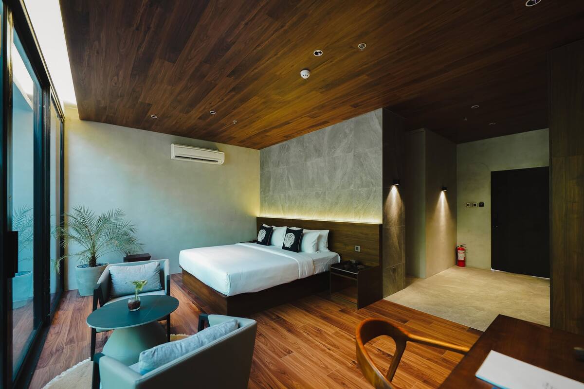 Een moderne hotelkamer met een houten plafond, een bed met witte lakens, een klein rond tafeltje, een bank en een plant in pot.