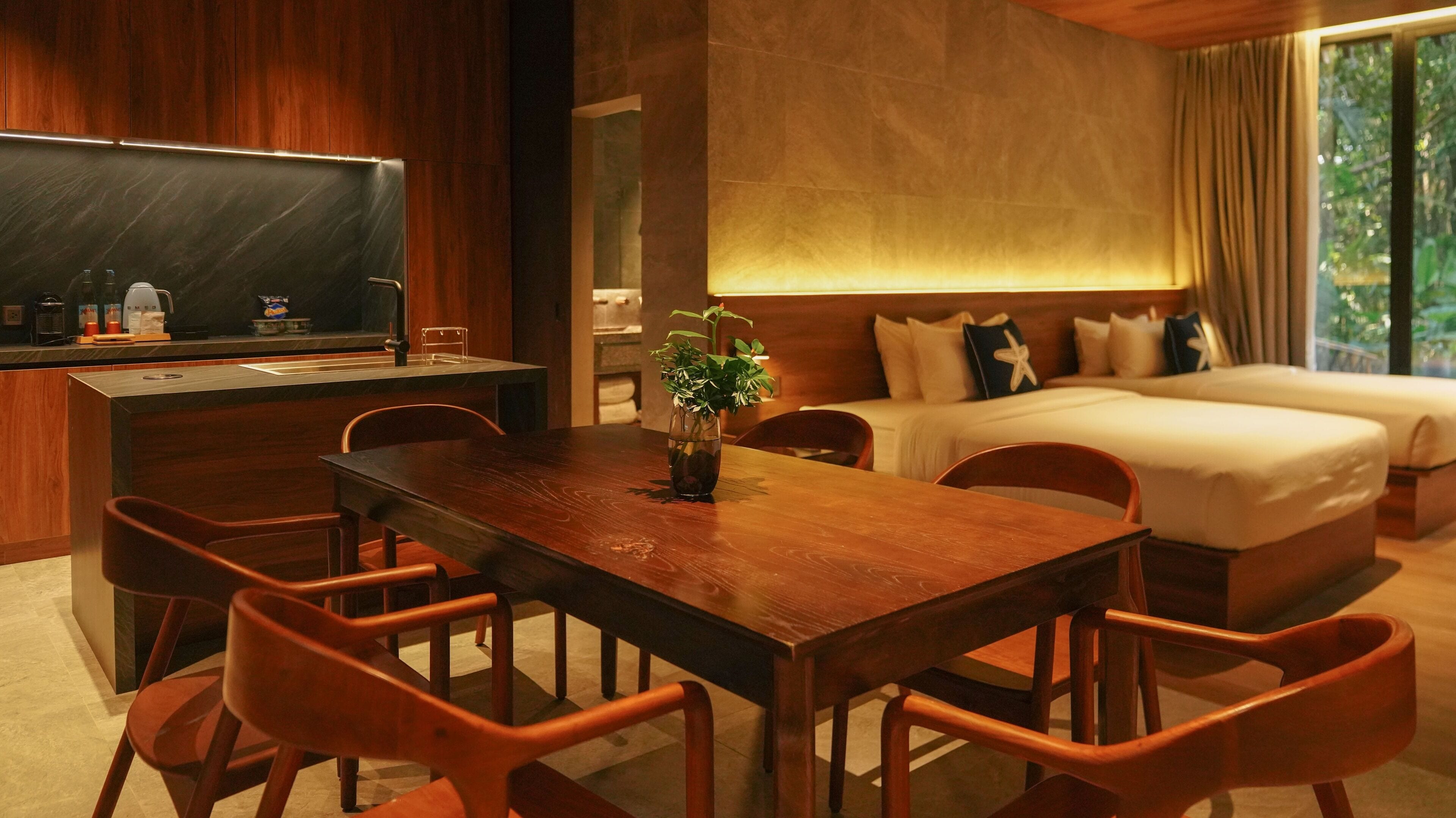 Une chambre d’hôtel avec une table à manger en bois, des chaises, un lit avec deux oreillers, une table de chevet et une fenêtre avec des rideaux.