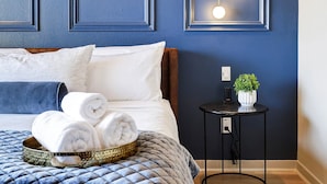Ein ordentlich bezogenes Bett mit blauer Bettdecke, ein Nachttisch mit Pflanze und eine Wandleuchte.