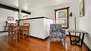 Une pièce comprenant une table et des chaises en bois, un fauteuil à motifs, une petite table avec une lampe et une fenêtre avec des stores.