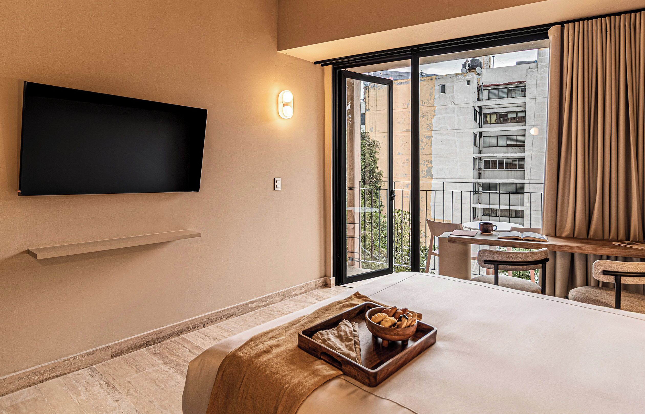 A hotel room with a flat-screen TV, a dining area with a table and chairs, a large window with curtains, and a small balcony.