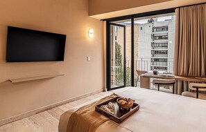 Een hotelkamer met een flatscreen-tv, een eethoek met tafel en stoelen, een groot raam met gordijnen en een klein balkon.