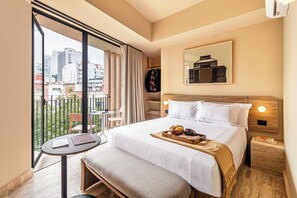 Camera d'albergo con un letto grande, un tavolino con un vassoio di cibo, una panca, una sedia, un balcone con vista sulla città e un'opera d'arte appesa al muro.