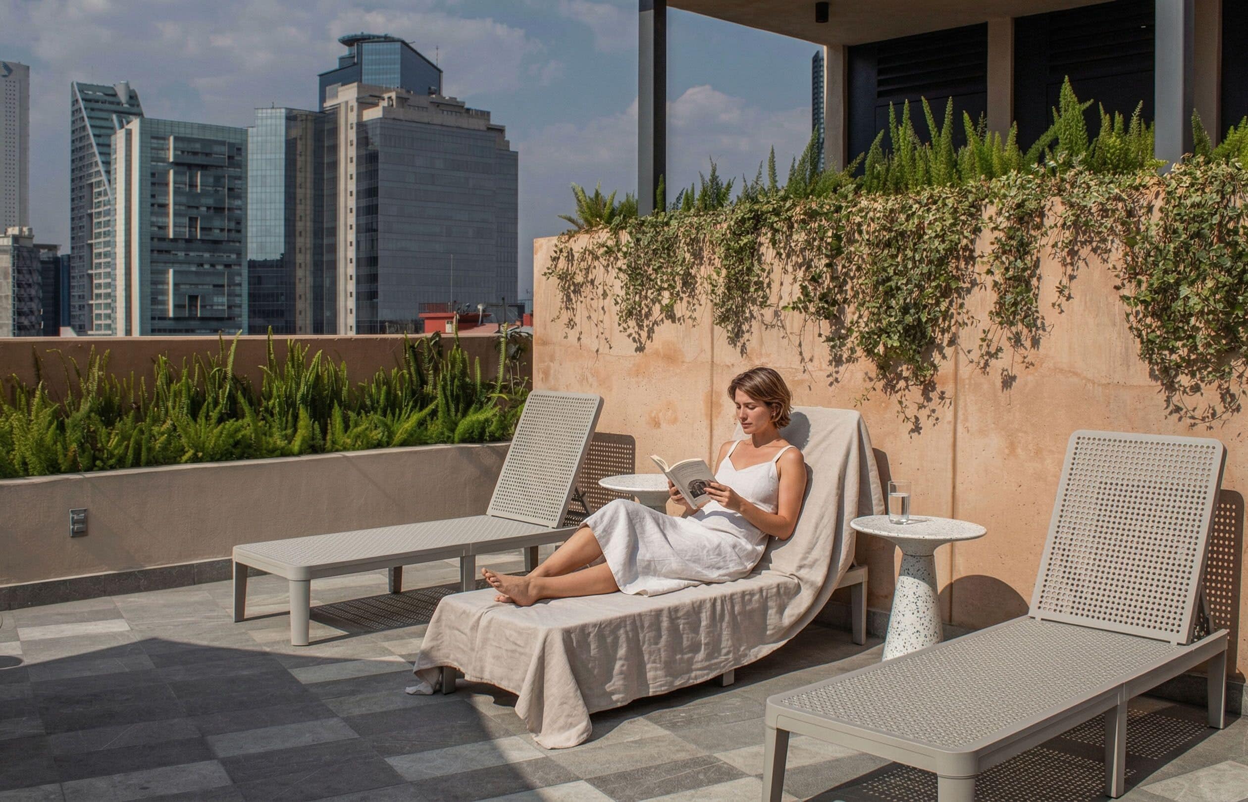 Un salón en la azotea con vista a la ciudad, una silla blanca para lounge, una mesita pequeña y una persona leyendo un libro.