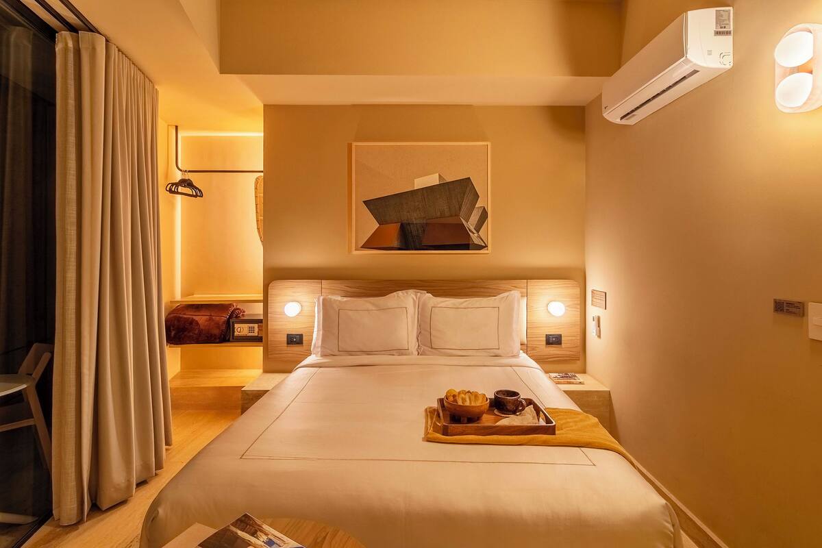 A hotel room with a bed, bedside tables, a wall-mounted air conditioner, and a tray with breakfast items.