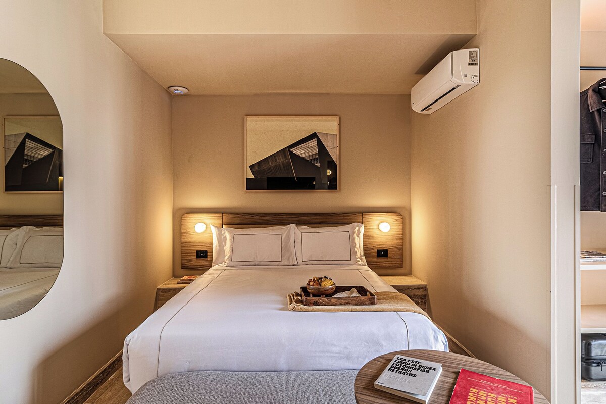 Habitación de hotel con cama, mesitas de noche, aire acondicionado de pared, un cuadro enmarco y un espejo.