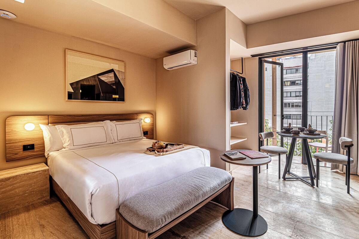 A hotel room with a bed, bedside table, bench, and a balcony with a table and chairs.