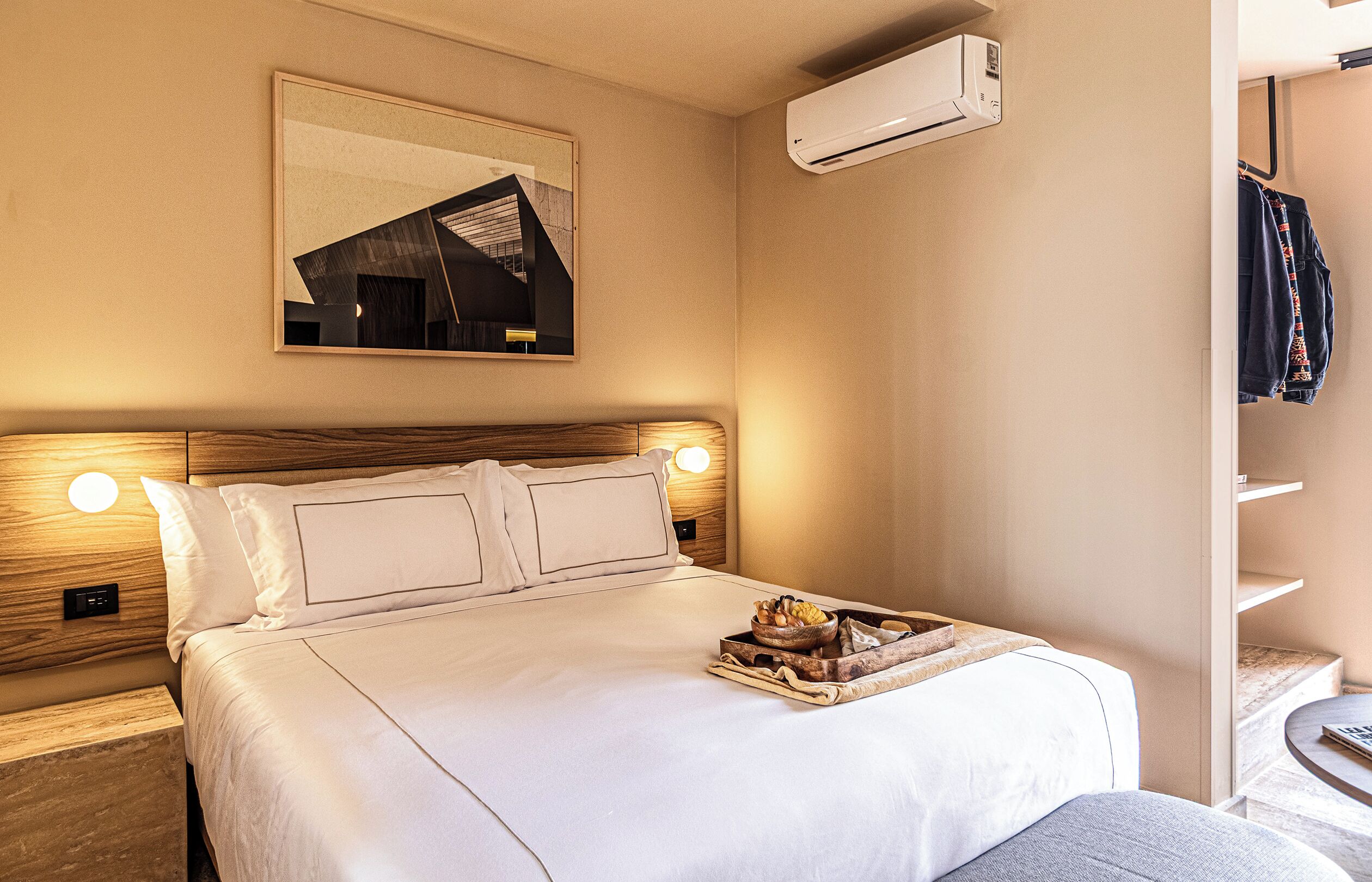 A hotel room with a bed, bedside lamps, a wall-mounted air conditioner, a framed picture, and a clothing rack.