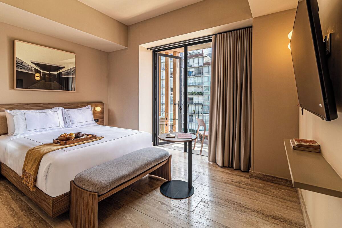 A hotel room with a bed, a bedside table, a bench, a TV, a window with curtains, and a view of buildings outside.