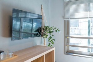 A room with a wooden console table, a mounted TV, a hanging plant, and a window with blinds. - Mi-HOTEL OTSUKA (Tokyo)