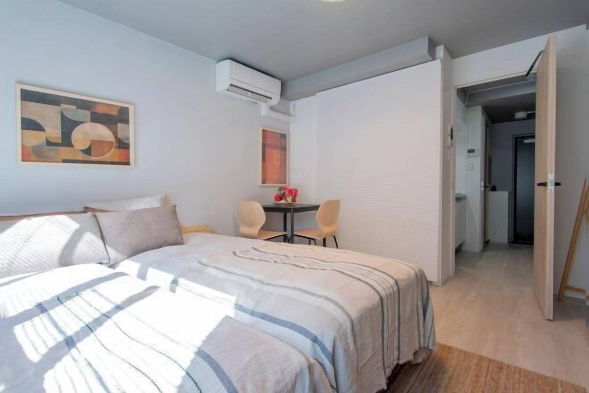 A bedroom with a bed, a desk, and chairs, an air conditioning unit, and a hallway.