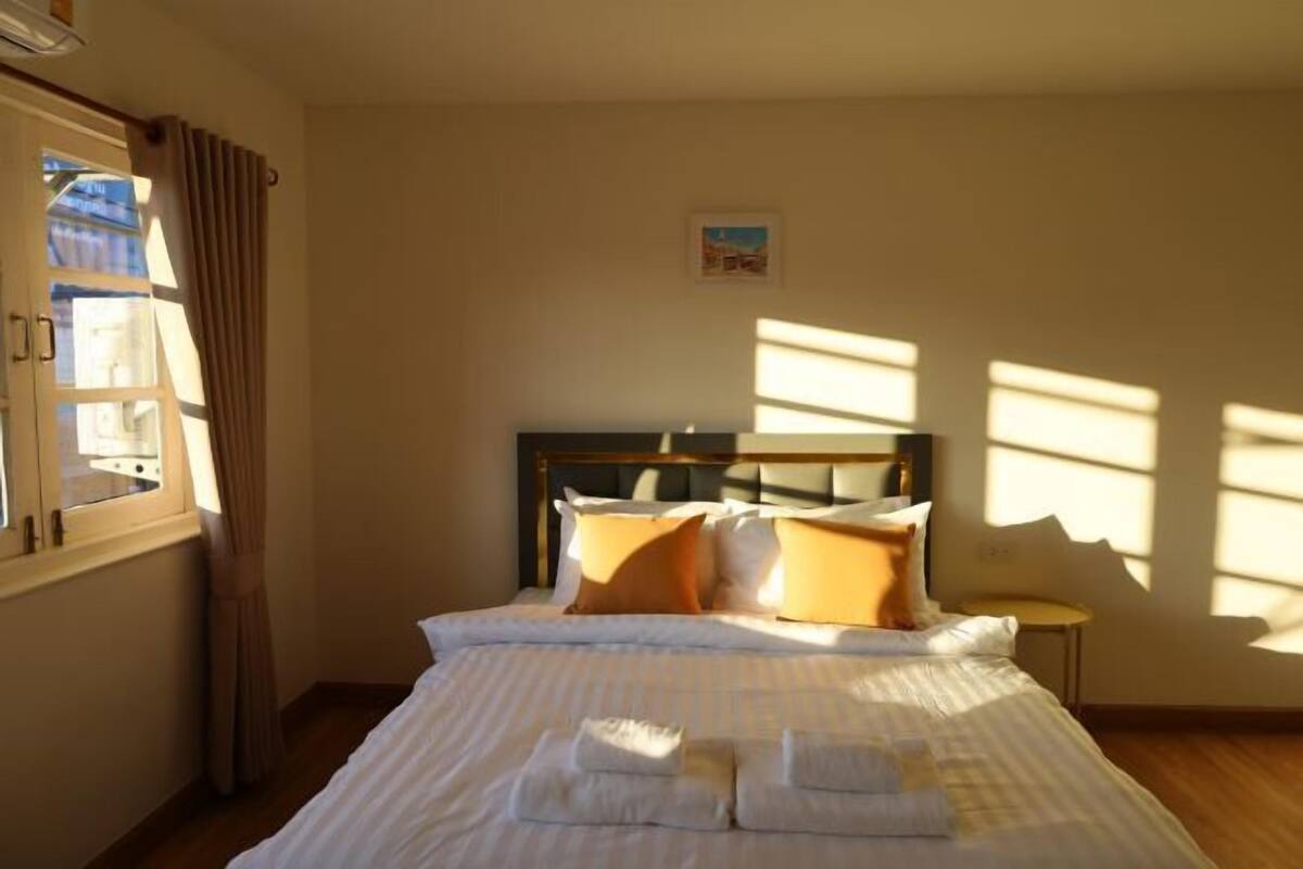 Uma cama bem arrumada com almofadas e um cobertor, uma mesinha de cabeceira, uma janela com cortinas e um pequeno quadro na parede.