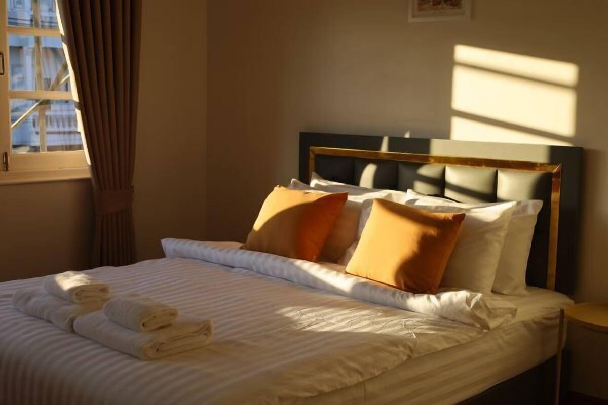 A neatly made bed with white linens and a headboard, a window with curtains, and a small bedside table.