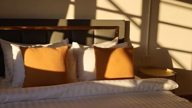 A bed with white bedding and multiple pillows, a bedside table, and a wall with a window casting shadows. - The 5ive Homestay (Chiang Rai)