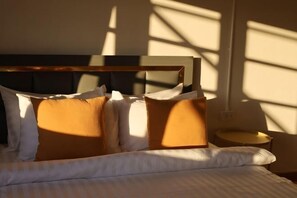 Ein Bett mit weißer Bettwäsche und mehreren Kissen, ein Nachttisch und eine Wand mit einem Fenster, das Schatten wirft.