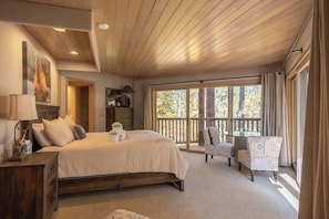 Ein Schlafzimmer mit einem großen Bett, einem Nachttisch, einer Lampe, einem Schminktisch, einem Sessel und einem Balkon mit Blick auf Bäume.