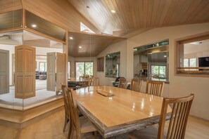 A spacious dining area with a long wooden table and chairs, a kitchen area with a large window, and a mirrored wall reflecting the room.
