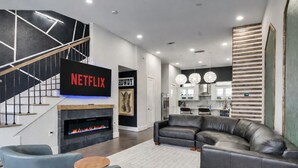 A modern living room with a fireplace, a staircase, a kitchen, and a flat-screen TV displaying 'NETFLIX'.