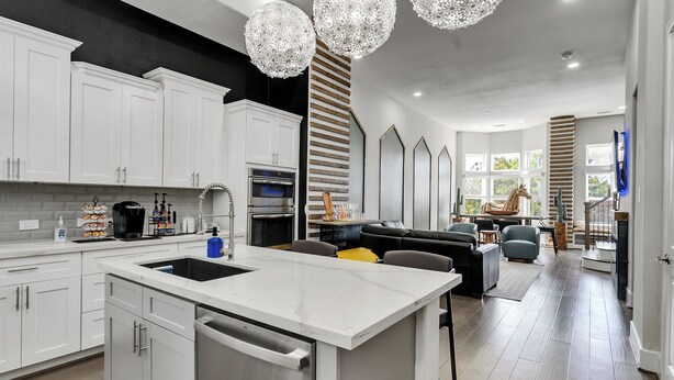 A modern kitchen with white cabinets, stainless steel appliances, and a central island. There are hanging crystal chandeliers and a large window providing natural light.