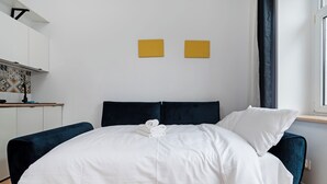 Une chambre moderne avec un grand lit, un canapé bleu foncé, un petit coin cuisine et deux œuvres d’art murales jaunes.