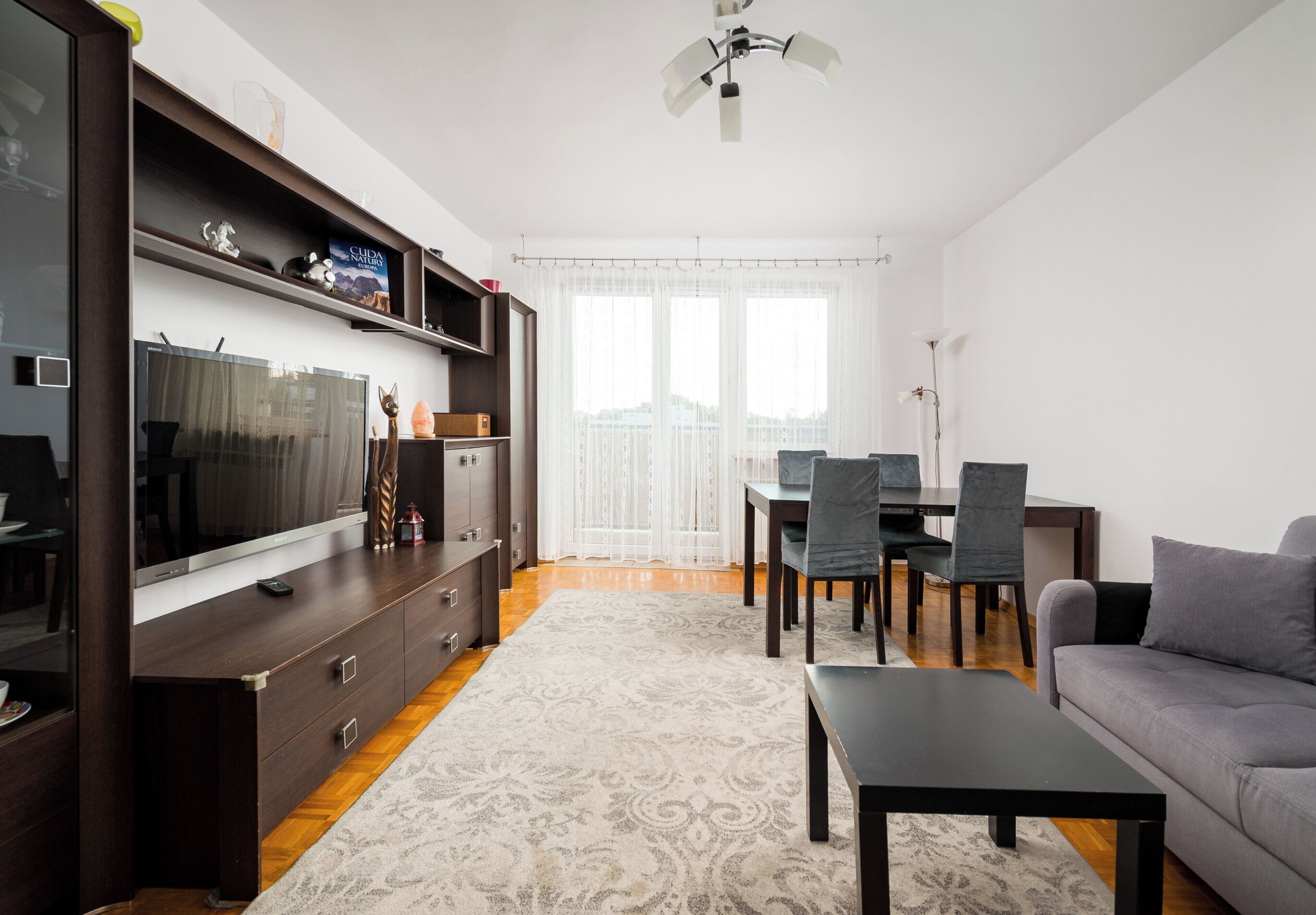 Un salon moderne doté d’un téléviseur à écran plat, d’un meuble de rangement en bois foncé, d’un canapé gris et d’un coin repas avec une table et des chaises.