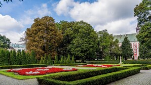 A formal garden with geometrically trimmed hedges and flower beds, a cobblestone pathway, and classical architecture in the background.
