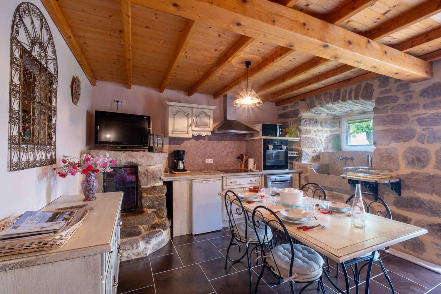 Una cocina acogedora con paredes de piedra, vigas de madera, una mesa de comedor con sillas y un televisor.