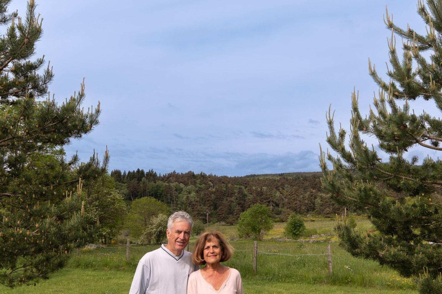 A couple standing in a grassy field with pine trees and a forest in the background.