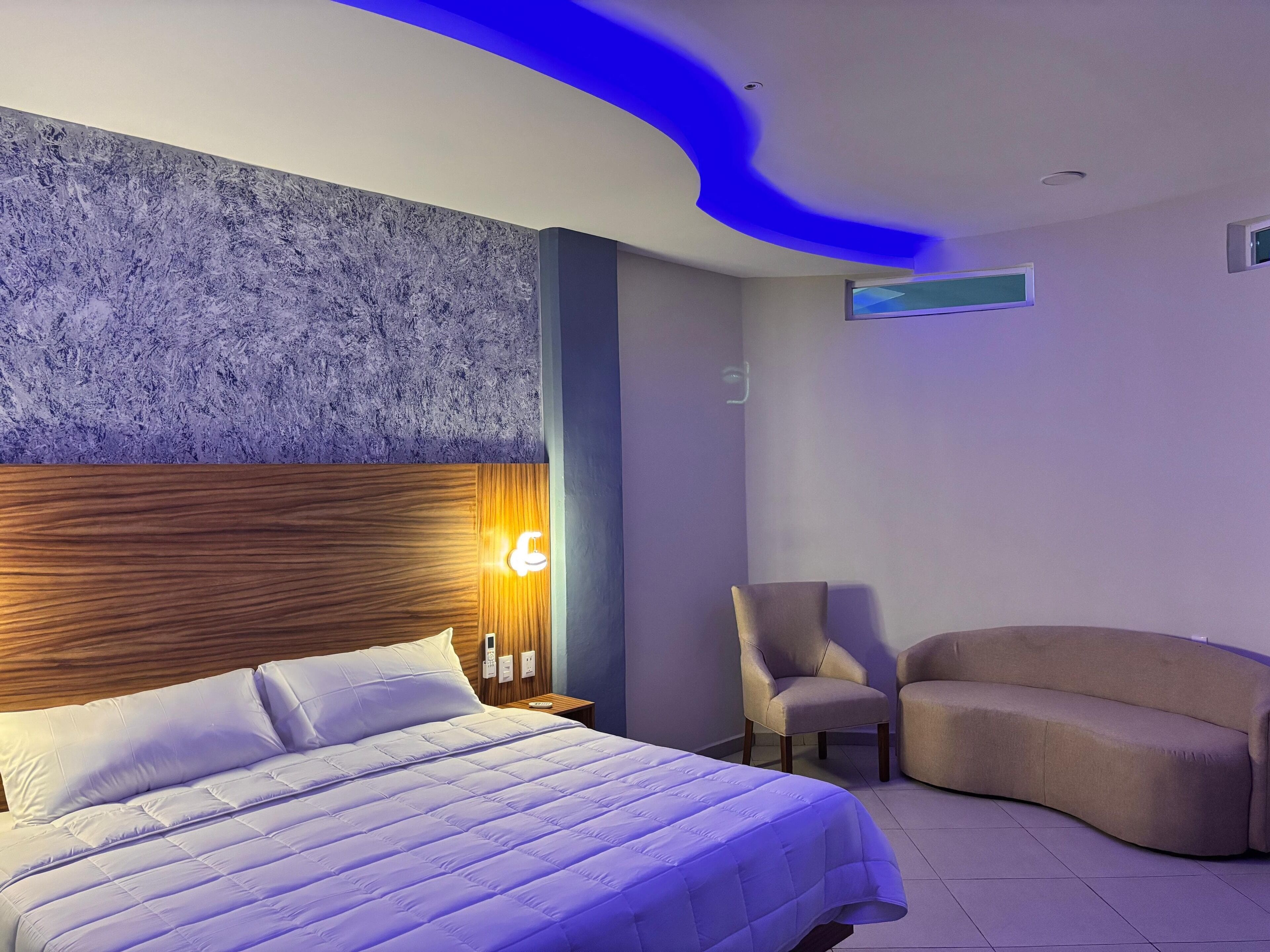 Una habitación de hotel moderna con una cama grande, cabecera de madera, una lámpara de pared, una mesita, dos sillones y un sofá curvo.