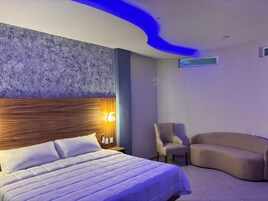 Una habitación de hotel moderna con una cama grande, cabecera de madera, una lámpara de pared, una mesita, dos sillones y un sofá curvo.