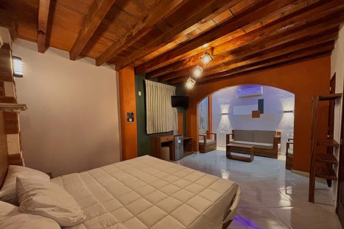A bedroom with a bed, a wooden ceiling, a small desk, and a view of a living area.