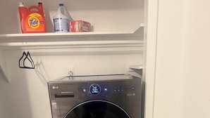 A compact laundry area with a front-loading LG washer and dryer, a striped laundry basket, and a shelf with cleaning supplies. - Emmanuel Midtown 2 BR 1.5 Bath Condo in Tulsa. Close to YMCA, Hospitals, Highway (Tulsa)