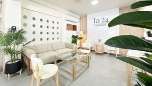 Un salon moderne avec un canapé beige, une table basse en verre et des chaises blanches. Un mur présente des découpes circulaires et un panneau indiquant « Vers 24 ».