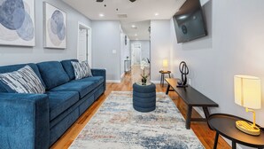 A modern living room with a blue sofa, a round ottoman, a wooden bench, a TV mounted on the wall, and a ceiling fan.