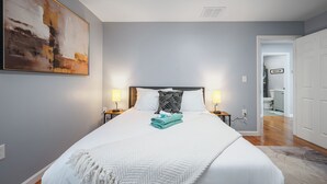A neatly made bed with a white comforter, a teal towel, and a white throw blanket. There are two bedside lamps, a large abstract painting on the wall, and a view into the bathroom through an open door.