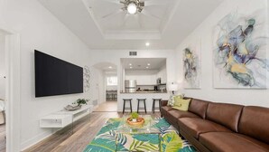 A modern living room with a brown leather sofa, a glass coffee table, and a colorful tropical rug. There is a flat-screen TV mounted on the wall, a kitchen area in the background, and abstract art on the wall. - Seven Eagles Springs: Near Disney, Golf, Free Water Park (Kissimmee)