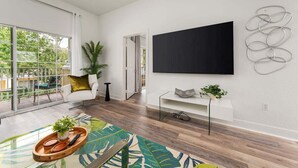 A modern living room with a large TV, wooden flooring, a glass coffee table, and a view of the outdoors. - Seven Eagles Springs: Near Disney, Golf, Free Water Park (Kissimmee)