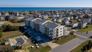 An aerial view of a coastal residential area with a prominent multi-story building featuring balconies and a parking lot. - Walk to Beach! Poolside Coastal Condo Retreat (Kill Devil Hills)