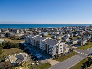 An aerial view of a coastal residential area with a prominent multi-story building featuring balconies and a parking lot.