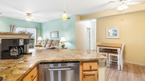 A kitchen with a dishwasher, a coffee maker, and a dining area with a table and chairs. - Walk to Beach! Poolside Coastal Condo Retreat (Kill Devil Hills)