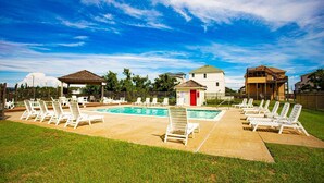 A pool area with lounge chairs, a small building with a red door, and a fenced yard. - Walk to Beach! Poolside Coastal Condo Retreat (Kill Devil Hills)