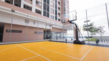 An indoor basketball court with a basketball hoop, surrounded by a high fence, and adjacent to a building with multiple floors and balconies.