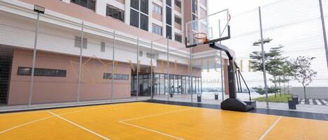 Una cancha de baloncesto cubierta con aro, rodeada por una valla alta, y contigua a un edificio con varios pisos y balcones.