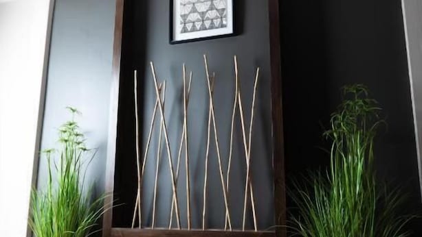 Ein Zimmer mit einer dunklen Wand, einem gerahmten geometrischen Kunstwerk, einer Holzablage mit Ästen und zwei Blumentöpfen.