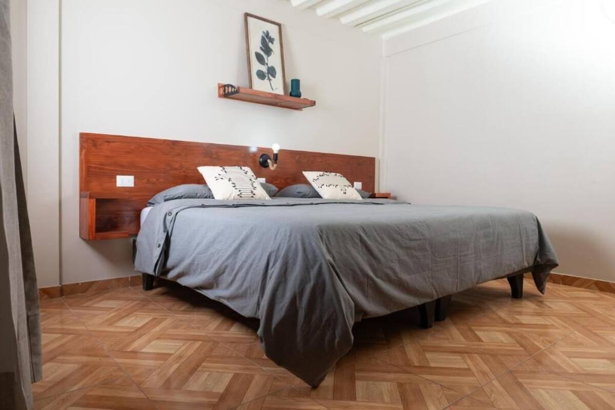 Un dormitorio con cabecera de madera, cama con ropa de cama gris, estante en la pared con una planta y suelo con patrón de espiga.