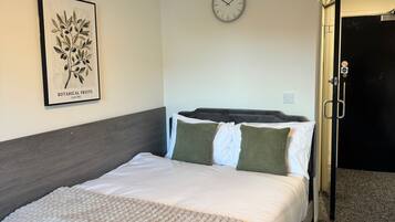A neatly made bed with a blue and beige quilt, a framed botanical print on the wall, a wall clock, and a door with a no-smoking sign.
