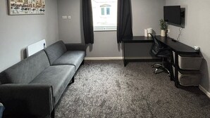 A small, furnished room with a gray sofa, a desk with a computer, a TV mounted on the wall, and a window with black curtains. - 248 Halls (Middlesbrough)