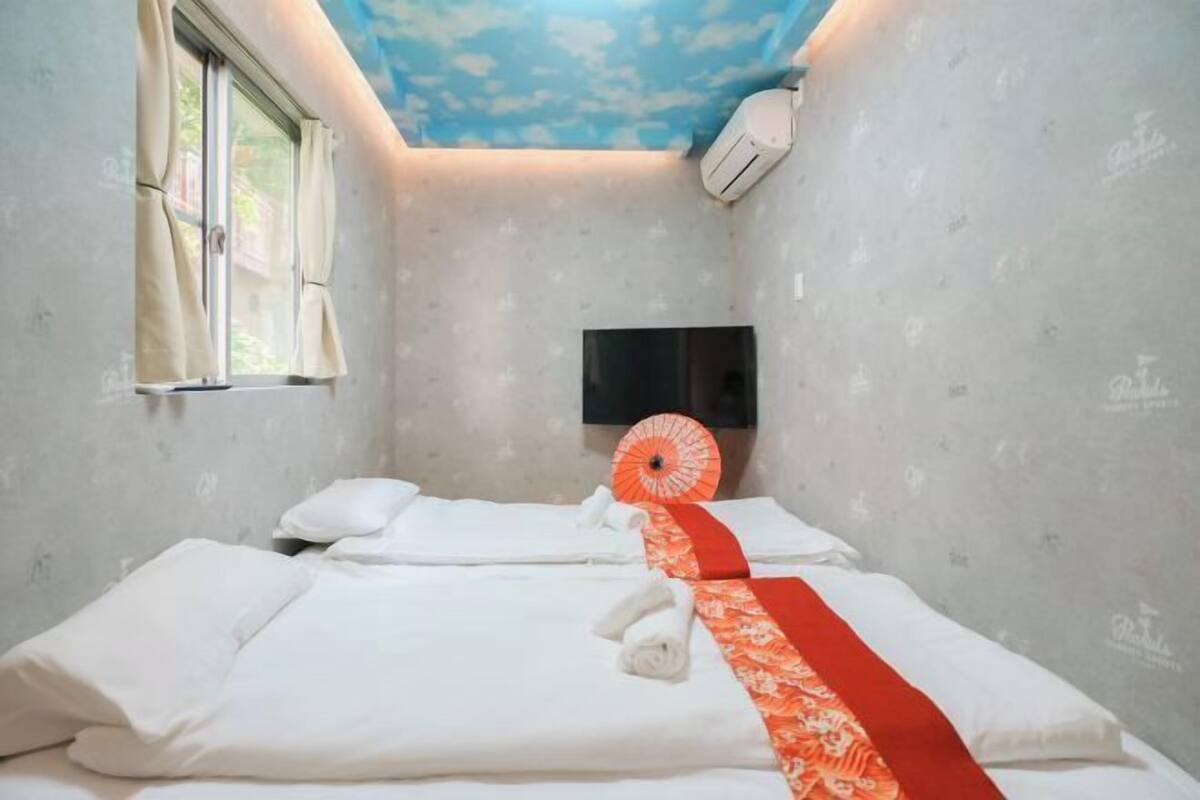 Ein Hotelzimmer mit zwei Betten, einem wandmontierten Flachbildfernseher und einem Fenster mit Vorhängen.