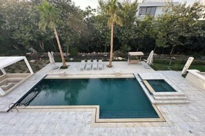 Une piscine rectangulaire, bordée d’une terrasse carrelée et entourée de chaises longues et de palmiers.