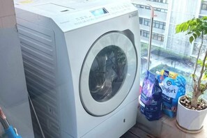 A front-loading washing machine with a control panel, a potted plant, and laundry detergent bags. - Vinhomes Metropolis Apartment - Comfort Stay Ha Noi (Hanoi)